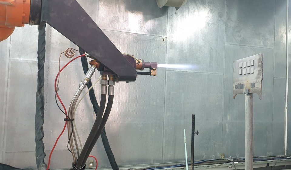 A thermal-spray coating cell in operation: a spray torch mounted on a robot arm, directed at a component.