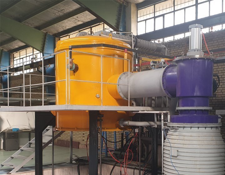 The furnace installation in its workshop hall: a large yellow furnace body on an elevated platform, connected by grey ducting to auxiliary vessels, under a steel roof.