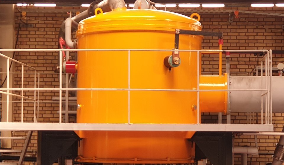 The vacuum heat-treatment furnace as built: an orange cylindrical vessel with a domed lid on a white steel platform with guardrails, inside a brick-walled workshop.